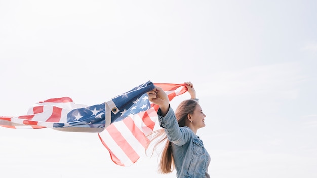 Gratis foto vrouw die en brede wuivende amerikaanse vlag hoog in hemel glimlacht houdt