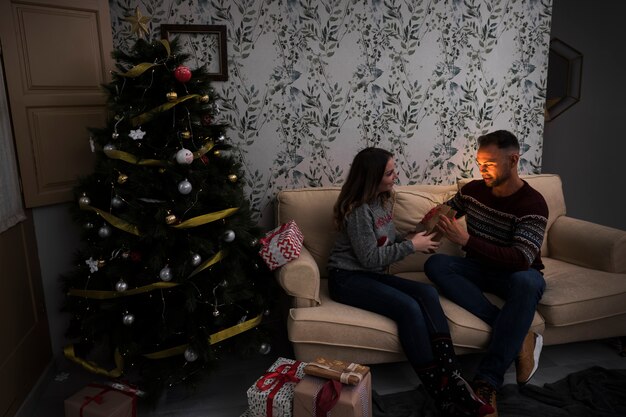 Vrouw die giftpakket voorstelt aan de mens op sofa dichtbij Kerstboom in ruimte