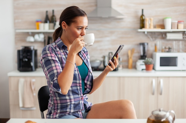 Vrouw die hete groene thee drinkt in de keuken die 's ochtends op smartphone bladert