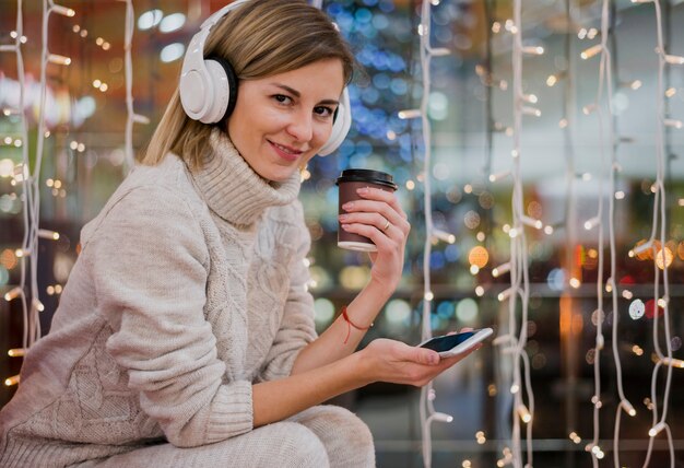 Vrouw die hoofdtelefoons draagt die kop en telefoon houden dichtbij Kerstmislichten