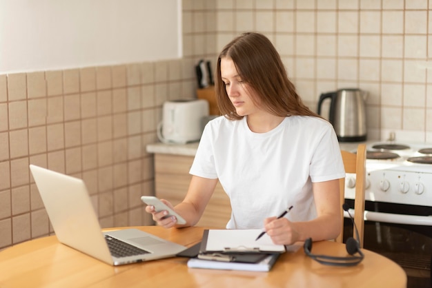 Vrouw die in de keuken werkt tijdens quarantaine met laptop en smartphone