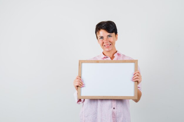 Vrouw die leeg frame in roze overhemd houdt en vrolijk, vooraanzicht kijkt.