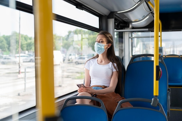 Vrouw die met de openbare bus reist en smartphone gebruikt terwijl ze een medisch masker draagt voor bescherming