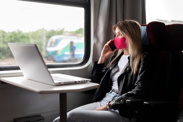 Vrouw die met de trein reist en aan de telefoon praat terwijl ze een medisch masker draagt en op een laptop werkt