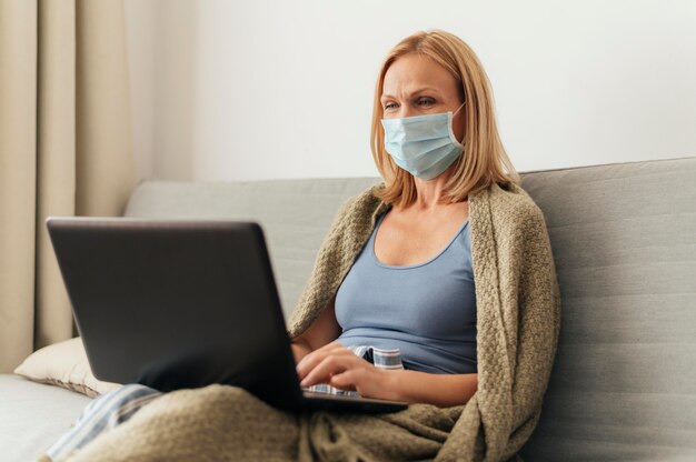 Vrouw die met medisch masker aan laptop thuis werkt tijdens zelfisolatie