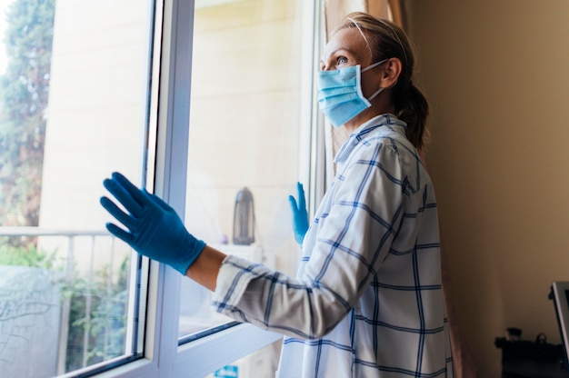 Vrouw die met medisch masker en handschoenen door venster kijkt tijdens quarantaine