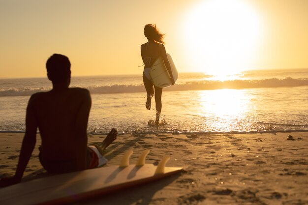 Vrouw die met surfplank loopt terwijl man het ontspannen op het strand tijdens zonsondergang