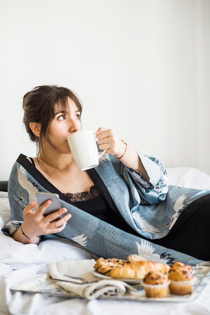 Gratis foto vrouw die op bed leunt die kop van koffie drinkt