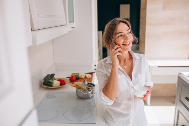 Vrouw die op de telefoon bij keuken en kokend ontbijt spreekt