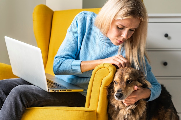 Vrouw die op laptop werkt vanuit fauteuil tijdens de pandemie en haar hond aaien