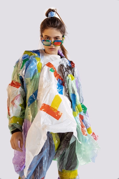 Vrouw die plastiek op witte muur draagt. Vrouwelijk model in kleding en schoenen gemaakt van afval. Mode, stijl, recycling, eco- en milieuconcept. Te veel vervuiling, we eten en nemen het op.