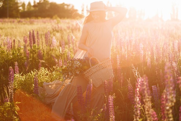 Vrouw die rieten zak in haar handen houdt die fedora hoed op zonsondergang op lupinegebied dragen
