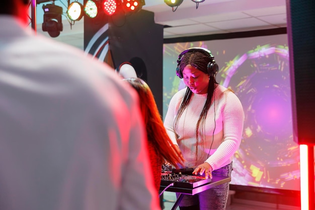 Vrouw dj in koptelefoon mixen van muziek op het podium tijdens elektronisch muziekconcert in nachtclub. Afro-Amerikaanse muzikant in koptelefoon die optreedt op discotheekfeest in club