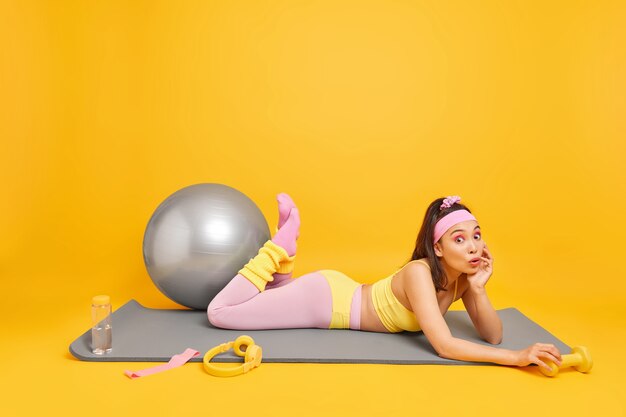 vrouw doet fysieke oefeningen op fitnessmat houdt halter gebruikt fitball voor pilatestraining gekleed in activewear