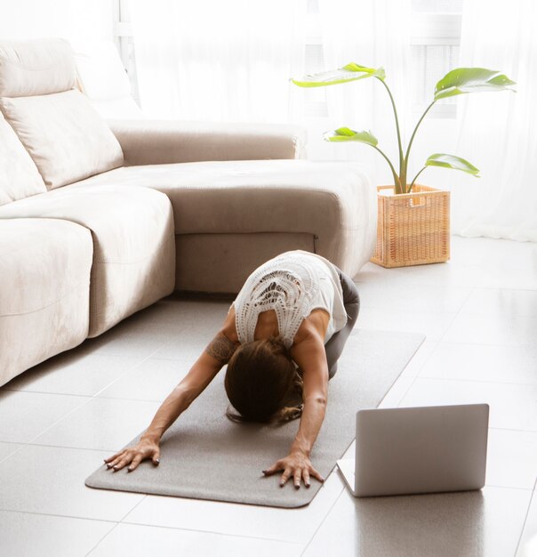 Vrouw doet yoga thuis op de mat met laptop op de vloer