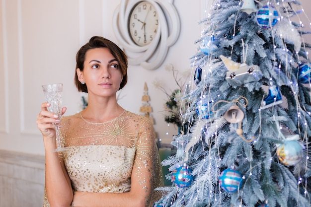 Vrouw draagt ?? glanzende gouden avond kerst jurk met glas champagne door bluenew jaar boom thuis