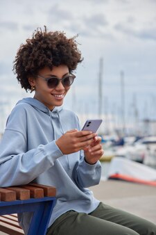 Vrouw draagt zonnebril en vrijetijdskleding gebruikt mobiele telefoon om online te chatten rust in de haven heeft vakantie op zee poseert in de haven heeft een vrolijke uitdrukking. lifestyle-concept