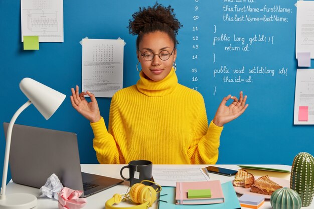 Vrouw freelance werknemer doet yoga-oefening op de werkplek, geniet van een rustige, rustige sfeer, draagt een ronde bril en trui