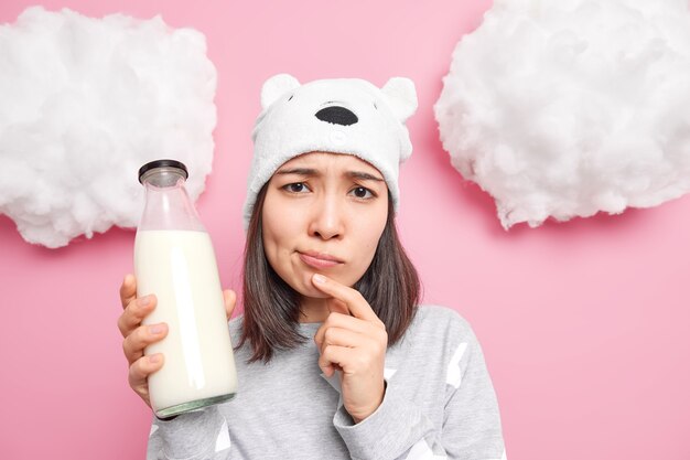 vrouw fronst gezicht houdt fles melk vast en denkt wat ze hiervan moet bereiden bij het ontbijt, gekleed in nachtkleding en hoed geïsoleerd op roze