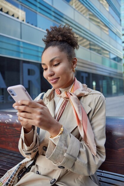 vrouw gebruikt smartphoneapparaat met draadloze internetchats online met volgers draagt beige jas en hoofddoek om nek zit op houten bankje in stedelijk gebied