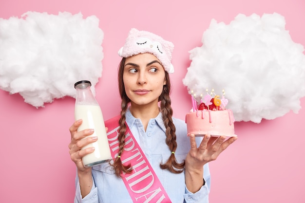 vrouw geconcentreerd opzij houdt heerlijke cake en fles melk die een feestelijk diner gaat hebben op haar verjaardag draagt een slaapmasker shirt geïsoleerd op roze