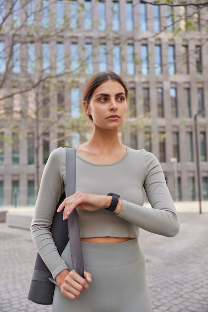 vrouw gekleed in activewear controleert de tijd op smartwatch draagt fitnessmat poses tegen modern stadsgebouw dat gaat trainen ziet er ergens bedachtzaam uit