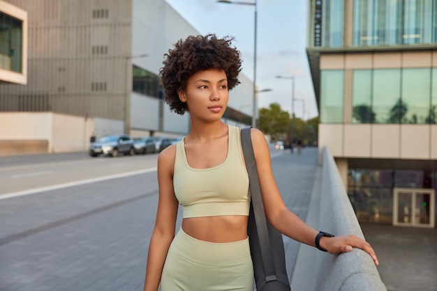 Gratis foto vrouw gekleed in trainingspak poseert op stedelijke weg draagt karemat gaat pilates-oefeningen doen met bank kijkt weg