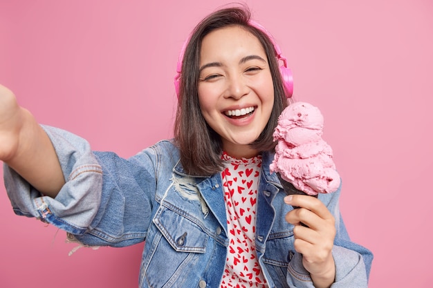 vrouw geniet van het eten van heerlijk ijsje in de zomer poseert voor selfie glimlacht breed luistert muziek via koptelefoon gekleed in spijkerjasje heeft plezier