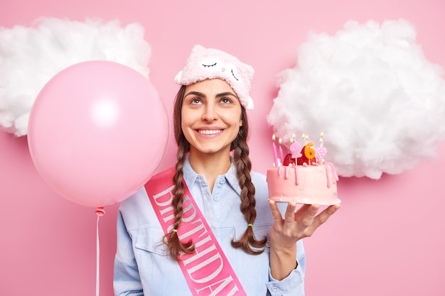 vrouw geniet van verjaardagsviering houdt heerlijke cake en opgeblazen ballon geconcentreerd boven, gekleed nonchalant drukt gelukkige emoties geïsoleerd op roze uit