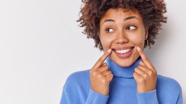 vrouw glimlacht breed wijst naar haar perfecte even tanden geeft aan dat op de hoeken van de lippen wegkijkt draagt casual blauwe trui geïsoleerd op wit met lege kopie ruimte.