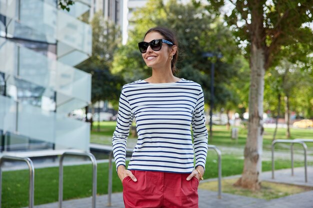 vrouw glimlacht vrolijk draagt zonnebril gestreepte trui en rode korte broek loopt door het stadspark geniet van zomerpromenade ademt frisse lucht staat in de buurt van stedelijke omgeving