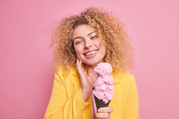 vrouw glimlacht zachtjes kantelt hoofd heeft vrolijke stemming eet smakelijk roze ijs tijdens warme zomerdag gekleed in modieuze gele jas poses binnen. Lekker dessert