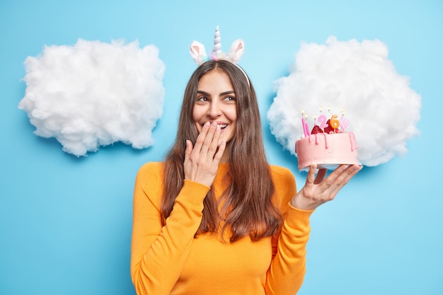 vrouw grinnikt vrolijk tegen mond met hand viert verjaardag houdt heerlijke feestelijke taart draagt casual oranje trui eenhoorn