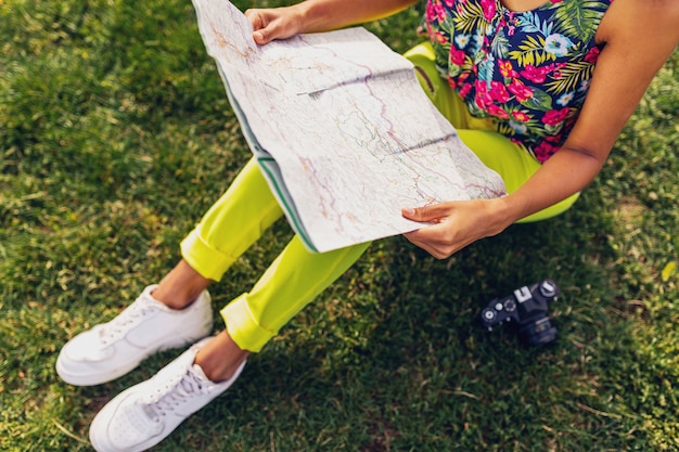 Vrouw handen met kaart, reiziger met camera plezier in park zomer mode stijl, kleurrijke hipster outfit, zittend op gras, gele broek