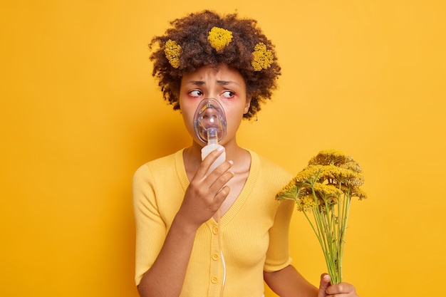 Gratis foto vrouw heeft astmatische aanval gezondheidsproblemen draagt een zuurstofmasker dat helpt om te ademen houdt wilde bloemen vast die een allergische reactie veroorzaken heeft rode opzwellende ogen geïsoleerd over gele muur