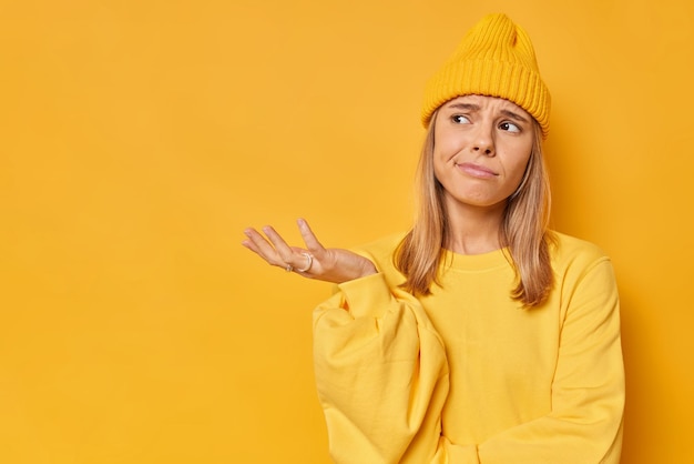 Gratis foto vrouw heft palm op haalt schouders op heeft besluiteloze uitdrukking gezichten moeilijke keuze draagt hoed en sweatshirt geïsoleerd op geel met lege kopie ruimte opzij