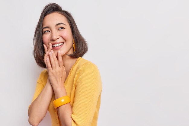 vrouw houdt handen bij elkaar glimlacht tandjes geconcentreerd weg met blije uitdrukking voelt erg blij draagt gele trui en armband poses op witte lege ruimte