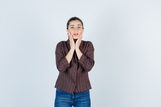 vrouw houdt handen op de wangen in shirt, spijkerbroek en kijkt verbijsterd, vooraanzicht.