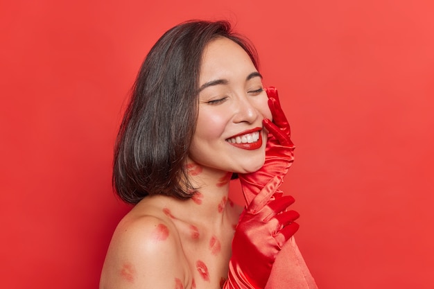 Gratis foto vrouw houdt ogen gesloten glimlacht aangenaam toont wit zelfs tanden raakt gezicht zachtjes poseert met blote schouders op rood voelt gelukkig