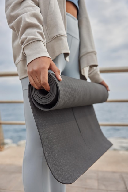 Vrouw houdt rubberen karemat vast voor yogabeoefening draagt sportkleding poseert buiten in de buurt van zee om te mediteren in goede fysieke conditie. welzijnsconcept