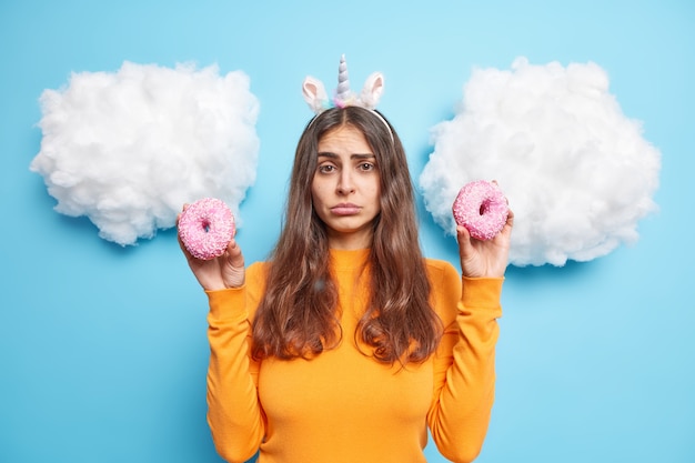 Gratis foto vrouw houdt twee geglazuurde donuts vast en heeft verleiding om junkmaaltijd te eten draagt oranje trui geïsoleerd op blauw