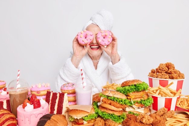 Vrouw houdt twee heerlijke geglazuurde donuts op ogen glimlacht blij omringd door junkfood verbruikt veel calorieën per dag draagt badjas