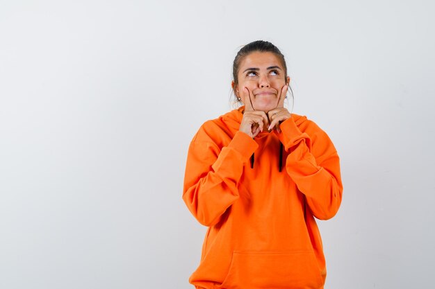 Vrouw houdt vingers op de wangen in oranje hoodie en ziet er schattig uit
