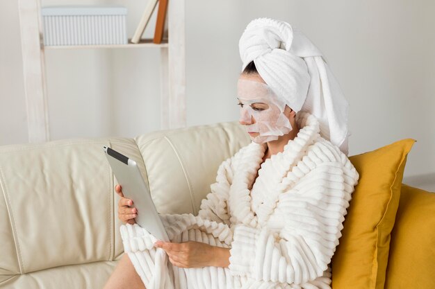 Vrouw in badjas met gezichtsmasker met haar tablet