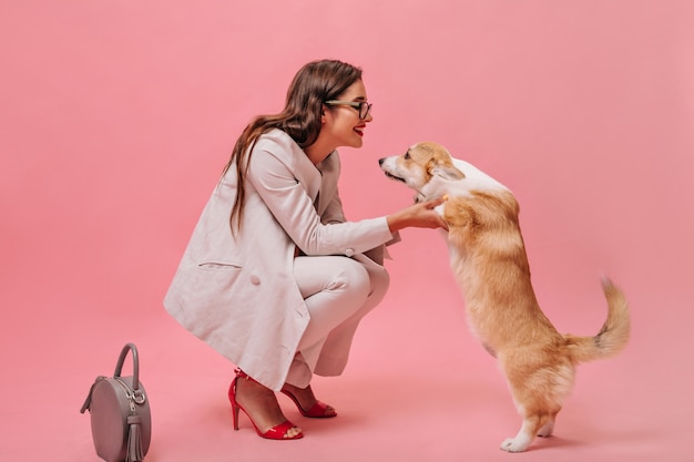 Vrouw in beige pak speelt met hond op roze achtergrond. Leuk mooi meisje met bril en rode hakken kijkt naar corgi en glimlacht.