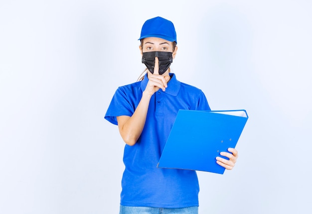 Vrouw in blauw uniform en zwart gezichtsmasker die een blauwe map vasthoudt en om stilte vraagt.