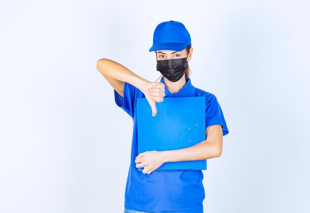 Vrouw in blauw uniform en zwart gezichtsmasker met een blauwe map en duim omlaag teken.