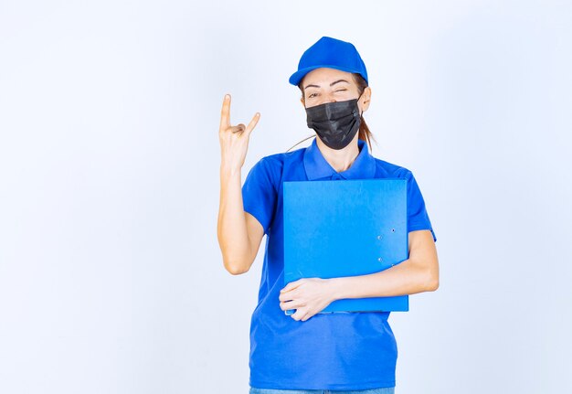 Vrouw in blauw uniform en zwart gezichtsmasker met een blauwe map en voelt zich positief en vriendelijk.