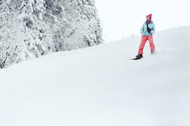 Vrouw in blauwe ski-jas en roze broek daalt de besneeuwde heuvel af op haar snowboard