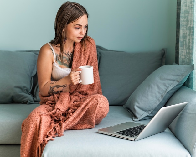 Vrouw in deken die koffie heeft en aan laptop thuis werkt tijdens de pandemie